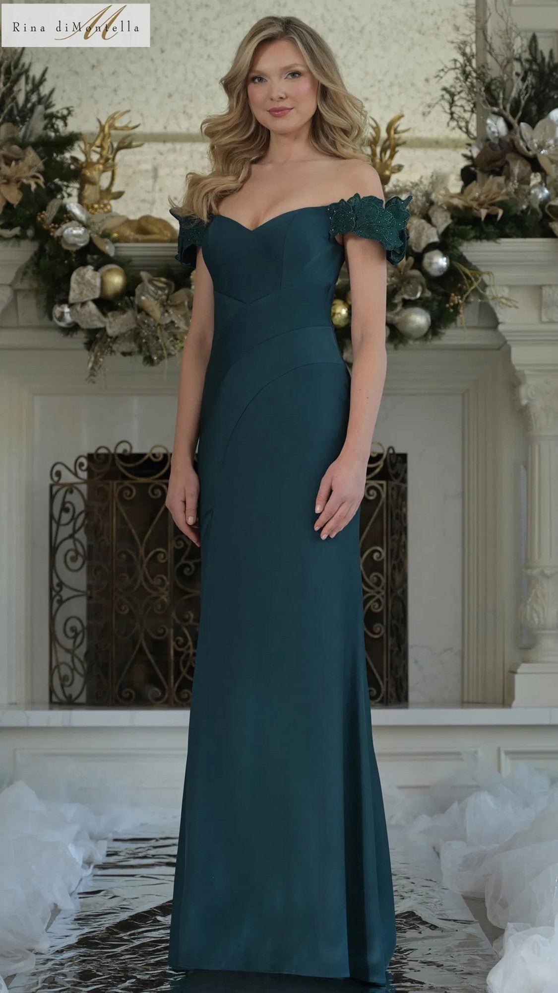 A woman stands indoors in a forest-colored Rina Di Montella RD3325 long formal dress with an off-the-shoulder design and lace trim, posed before a decorated white fireplace.